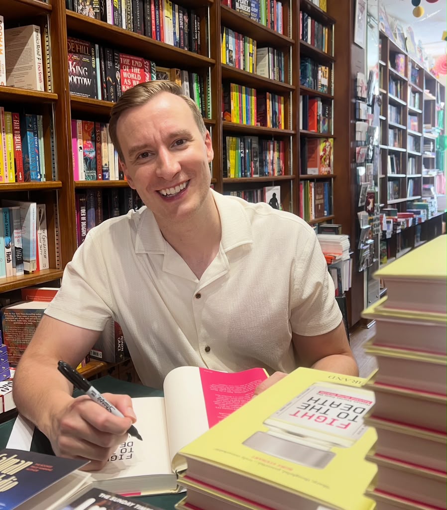Paul Brand smiling as he signs copies of Fight to the Death in a bookshop.